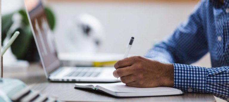 A person working at a laptop while holding a clipboard